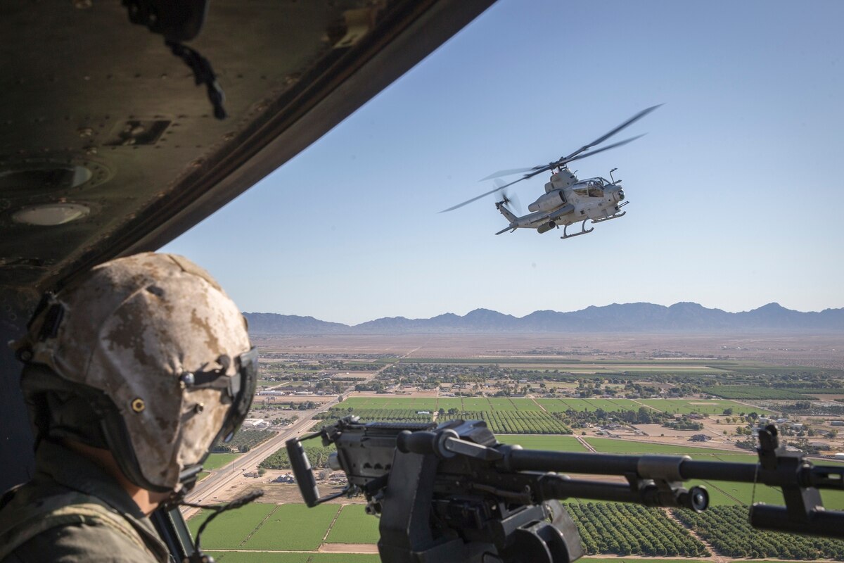 Picture of Marine AH-1Z Cobra and F-35B Lighting II flying over farmlands. Marine in the foreground with weapon